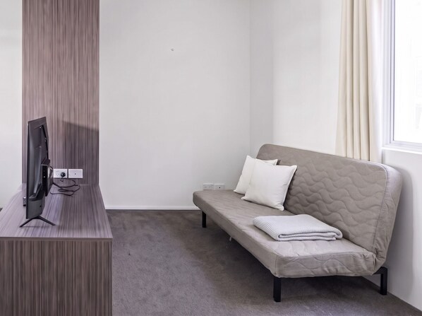 Una stanza con una TV a schermo piatto su un supporto in legno, un divano grigio con cuscini e una finestra con tende.