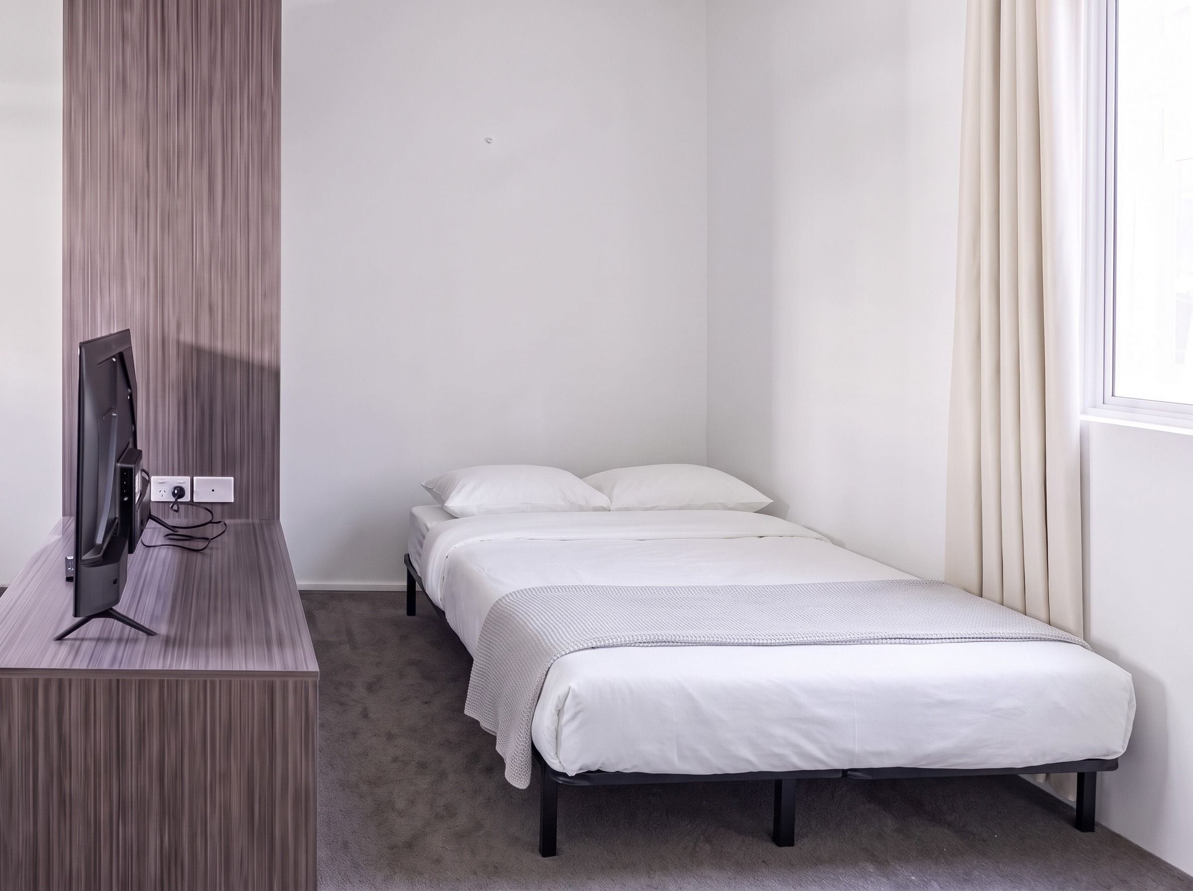 A small, single-bed room with a flat-screen TV on a wooden stand, a window with curtains, and a carpeted floor.
