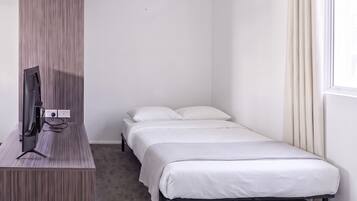 A small, single-bed room with a flat-screen TV on a wooden stand, a window with curtains, and a carpeted floor.