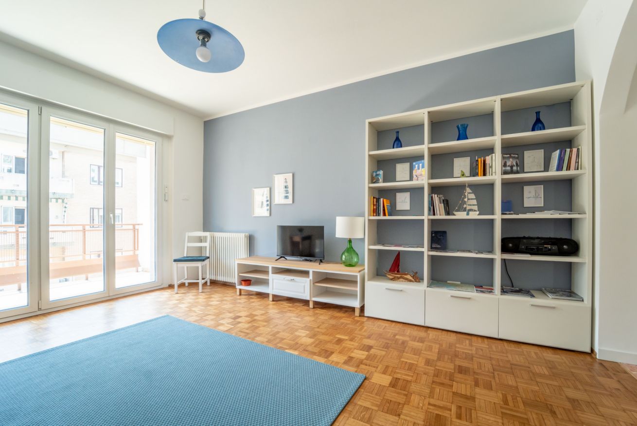 Ein Zimmer mit einem großen Fenster, einer blauen Matte, einem weißen Bücherregal und einem Fernseher.