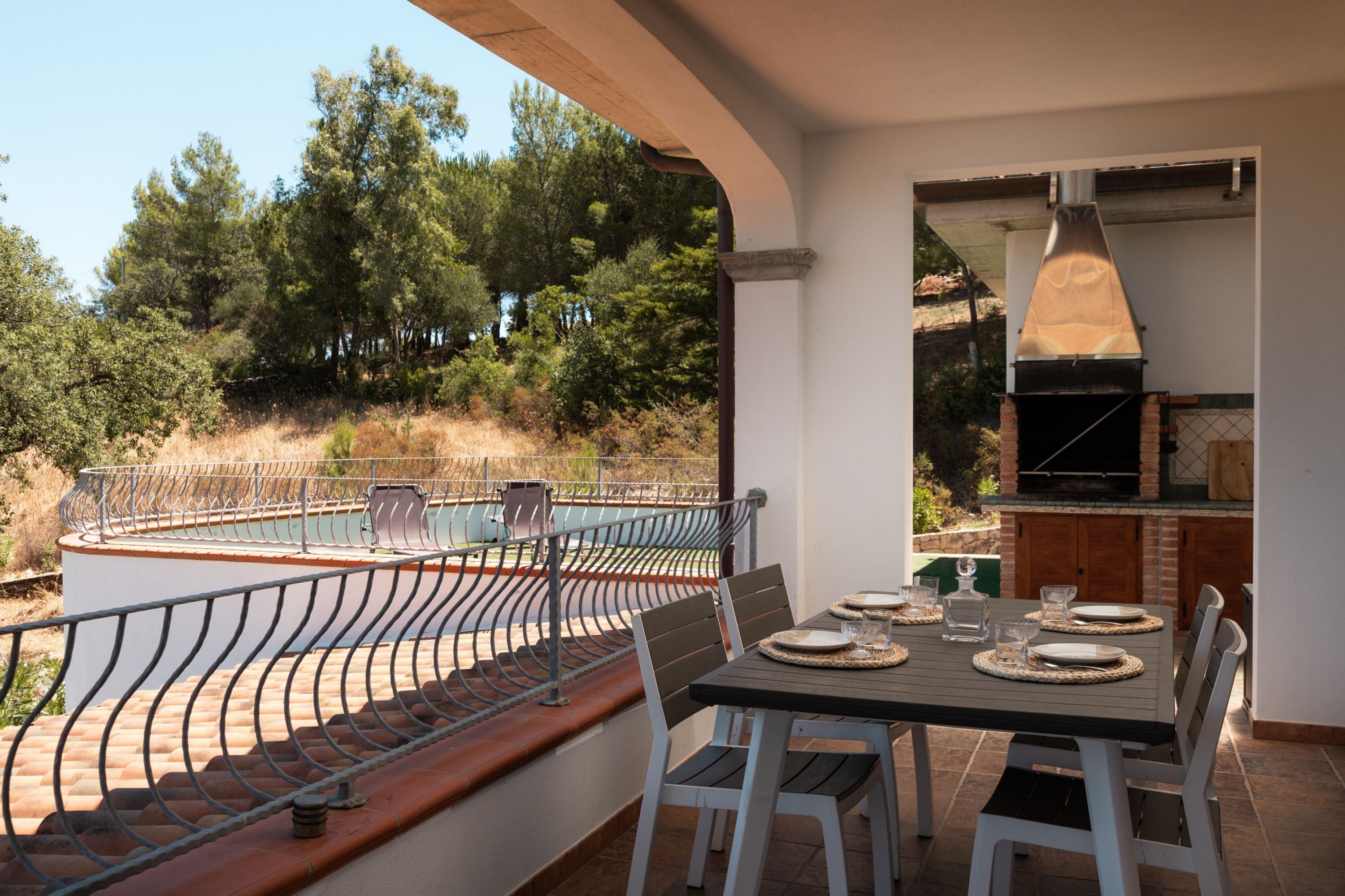 An outdoor dining area with a table set for four, overlooking a swimming pool and a forested hillside.