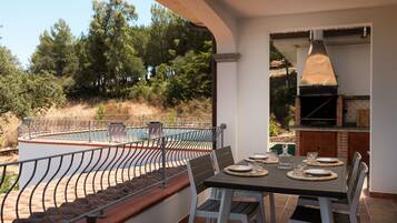 An outdoor dining area with a table set for four, overlooking a swimming pool and a forested hillside.