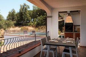 Uma área de refeições ao ar livre com uma mesa posta para quatro pessoas, com vista para uma piscina e um morro arborizado.