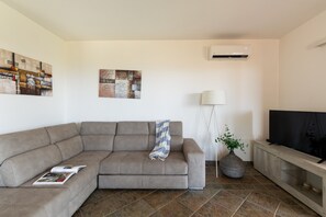 Sala de estar com sofá seccional, televisão de tela plana, luminária de chão, ar-condicionado fixado na parede e duas obras de arte na parede.