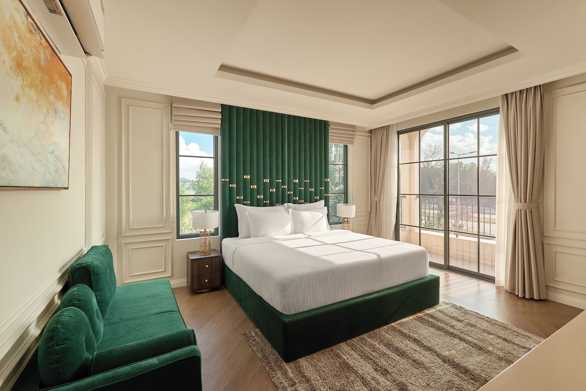A spacious bedroom with a large bed, a green accent wall, and a view of the outdoors through a large window.