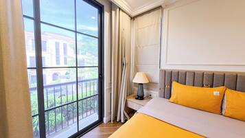 Ein Hotelzimmer mit großem Fenster, einem Bett mit gelber Bettwäsche, einem Nachttisch mit Lampe und Blick auf ein Gebäude sowie Grünflächen.