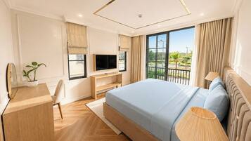 A modern hotel room with a large bed, a desk with a TV, a potted plant, and a view of a balcony.