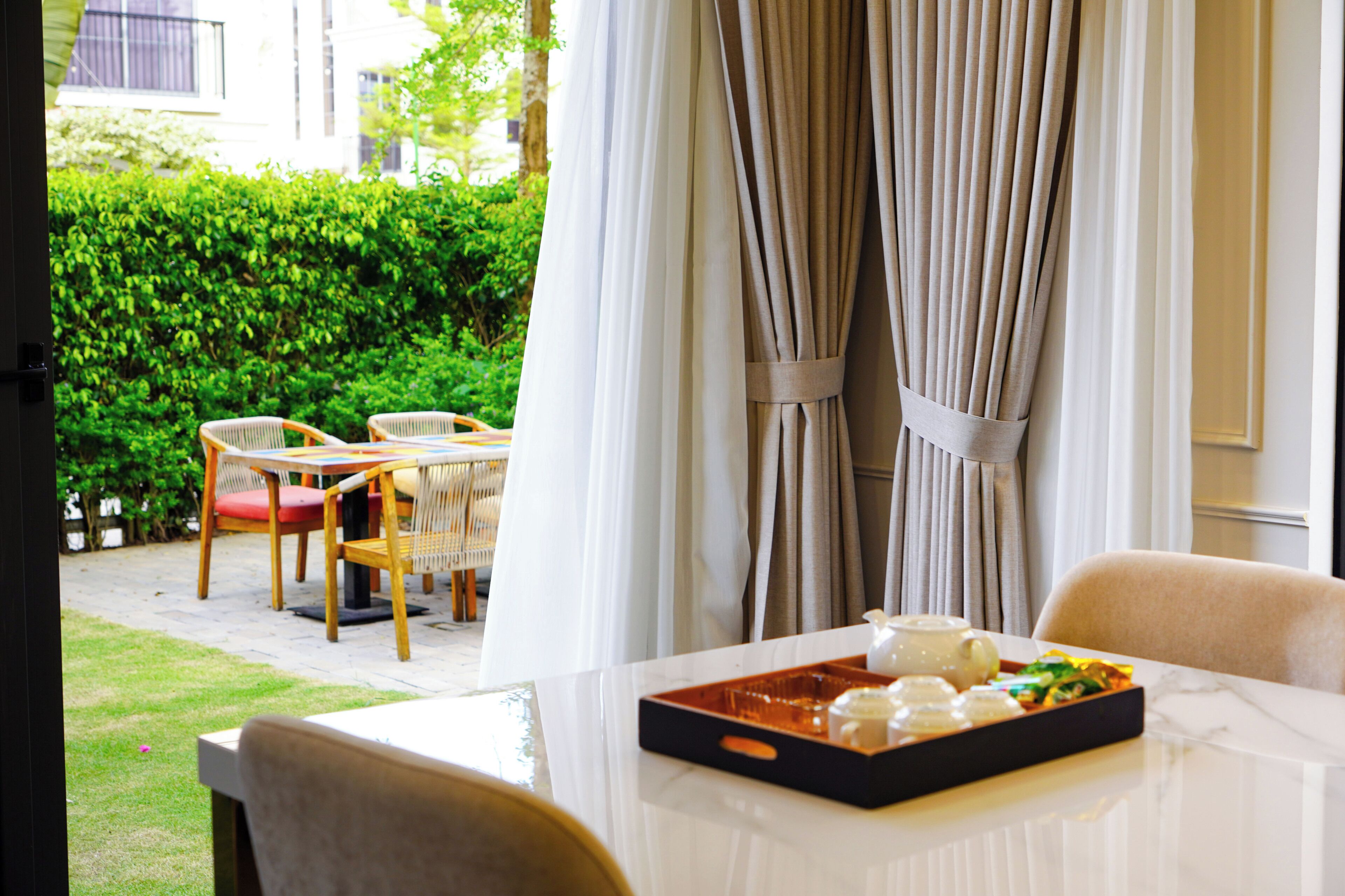 Un comedor con una mesa dispuesta para dos personas, una bandeja con comida y bebida, y vistas a un jardín con sillas y una mesa afuera.