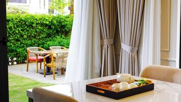 Un comedor con una mesa dispuesta para dos personas, una bandeja con comida y bebida, y vistas a un jardín con sillas y una mesa afuera.