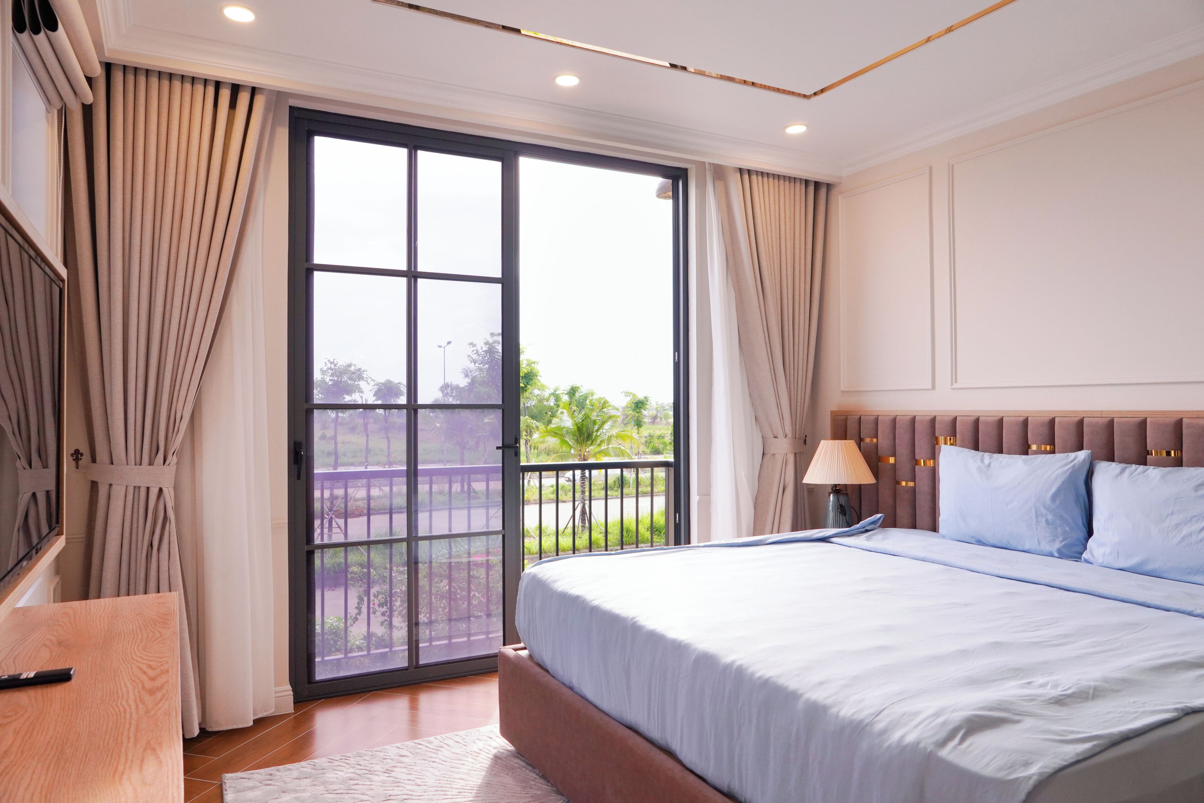 A hotel room with a large bed, a desk, a window with curtains, and a view of greenery outside.