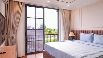 A hotel room with a large bed, a desk, a window with curtains, and a view of greenery outside.