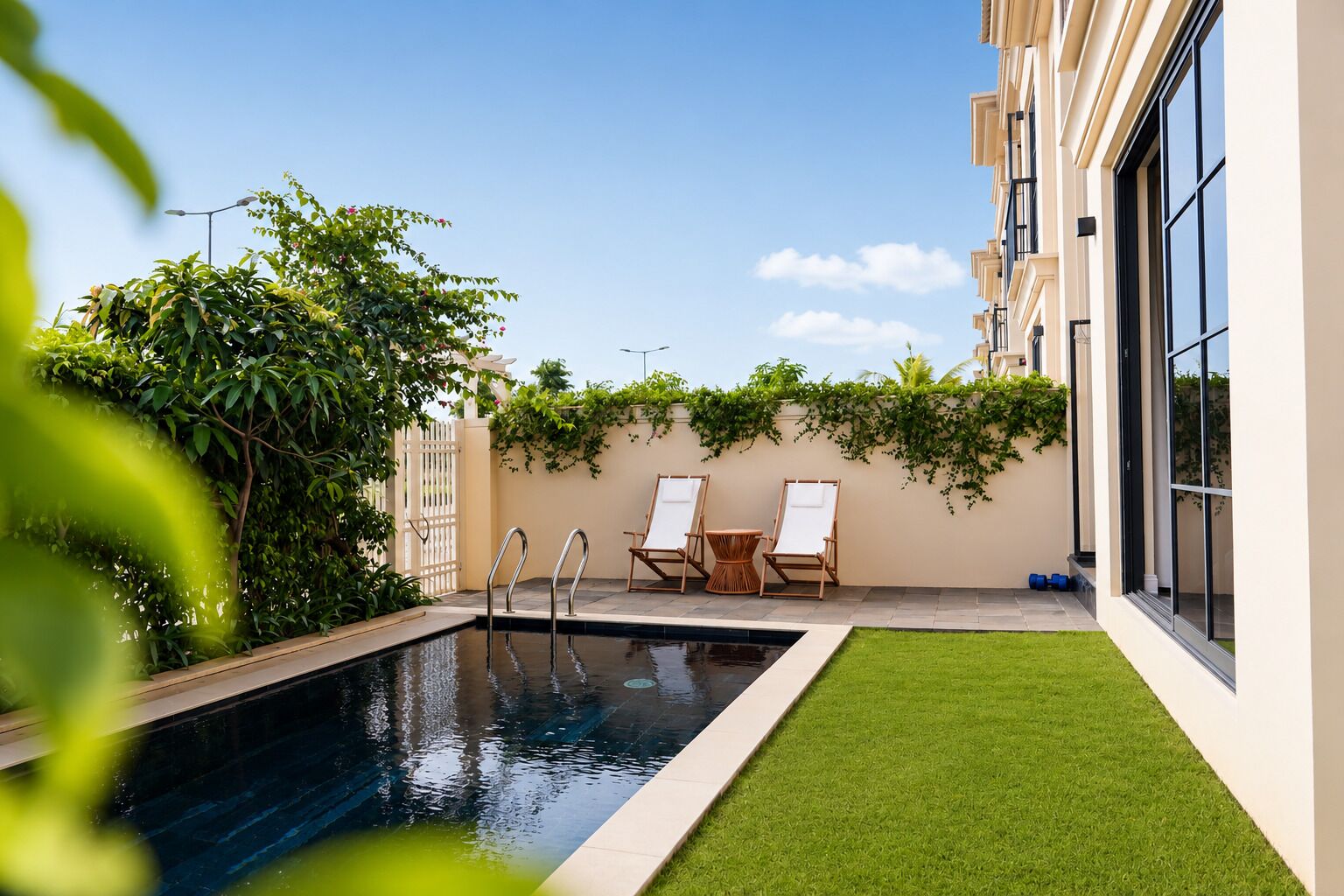 A swimming pool with two lounge chairs and a small table, surrounded by a well-maintained lawn and a building with large windows.