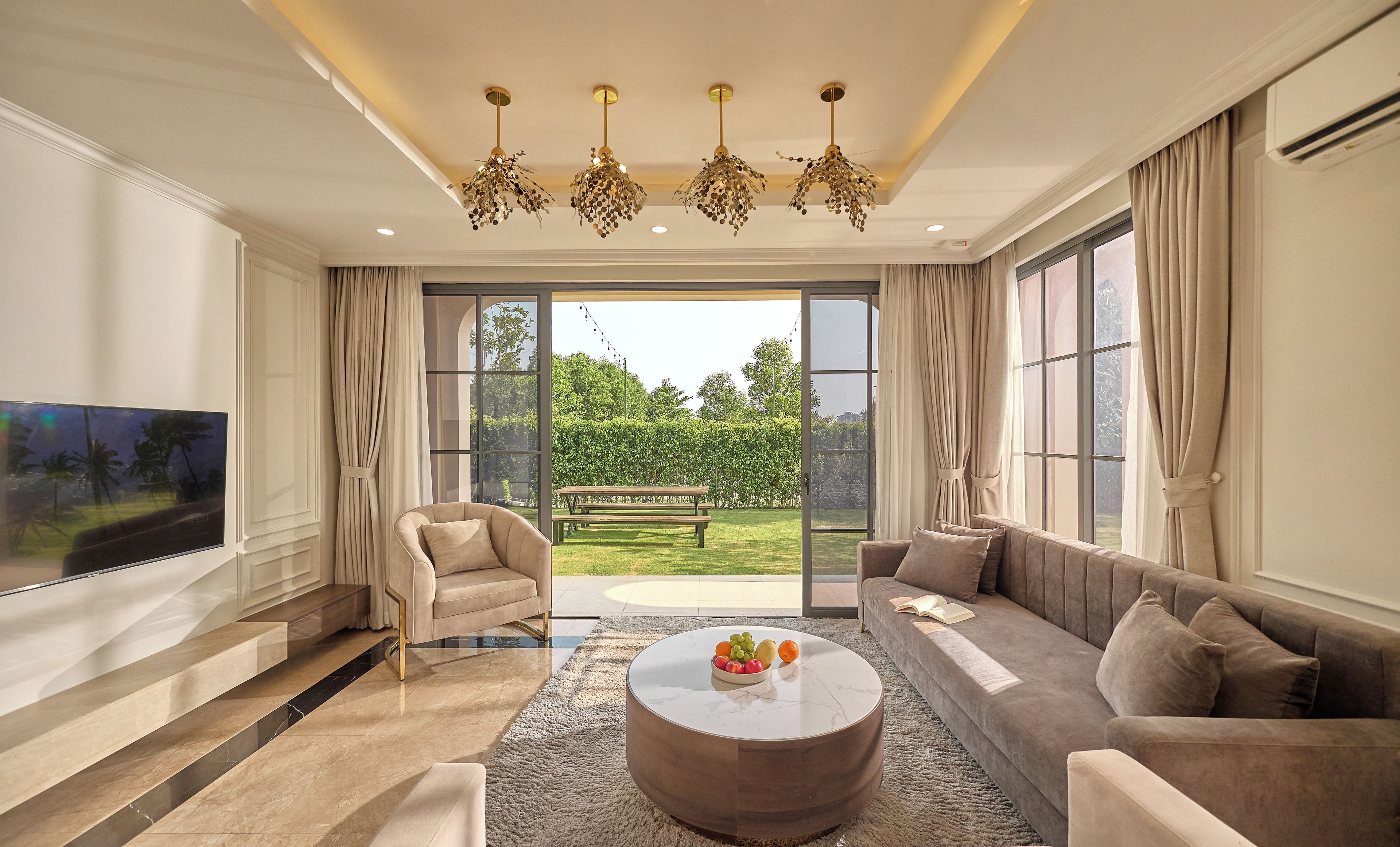 A spacious living room with a large flat-screen TV, a round coffee table with fruit, a sofa, and a chair.