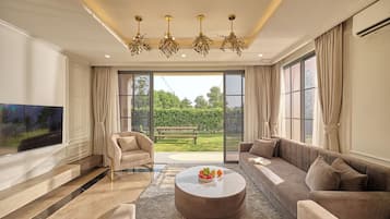 A spacious living room with a large flat-screen TV, a round coffee table with fruit, a sofa, and a chair.