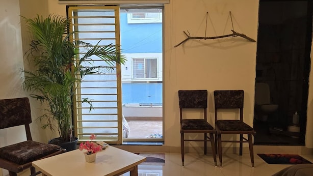 A living room with a table, two chairs, a potted plant, and a view through a sliding door. - The white Town Homestay (Indore)