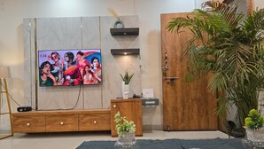 A modern living room with a flat-screen TV mounted on a marble wall, a wooden entertainment unit, a large potted plant, and a blue rug. - The white Town Homestay (Indore)