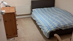 Una camera da letto con un letto, una cassettiera in legno, una scrivania con una lampada e un termosifone.