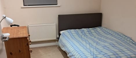 A bedroom with a bed, a wooden dresser, a desk with a lamp, and a radiator.