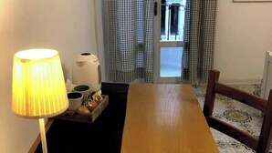 Una habitación con una mesa de madera, una lámpara, una bandeja con tazas y una tetera, y vistas a través de una ventana con cortinas a cuadros.