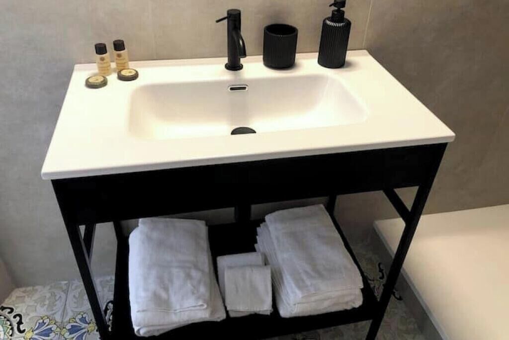 A bathroom with a white sink, black faucet, and two bottles of soap. There are towels stacked on a black shelf below the sink.
