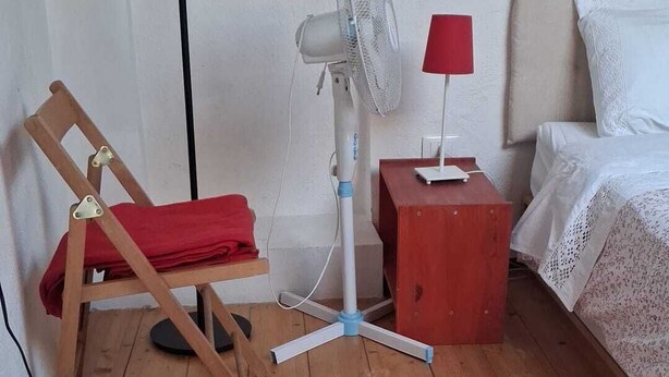 Ein Zimmer mit Holzboden, einem Standventilator, einem roten Stuhl, einem Nachttisch mit Lampe und einem Bett mit weißer Bettwäsche.