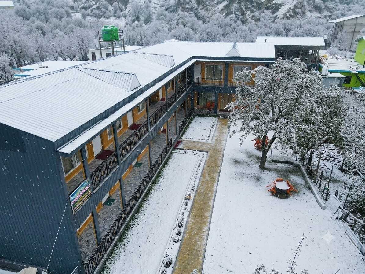 Un bâtiment dont le toit est recouvert de neige, doté d’un espace extérieur abrité avec des sièges, et offrant une vue sur des arbres et des montagnes également recouverts de neige.