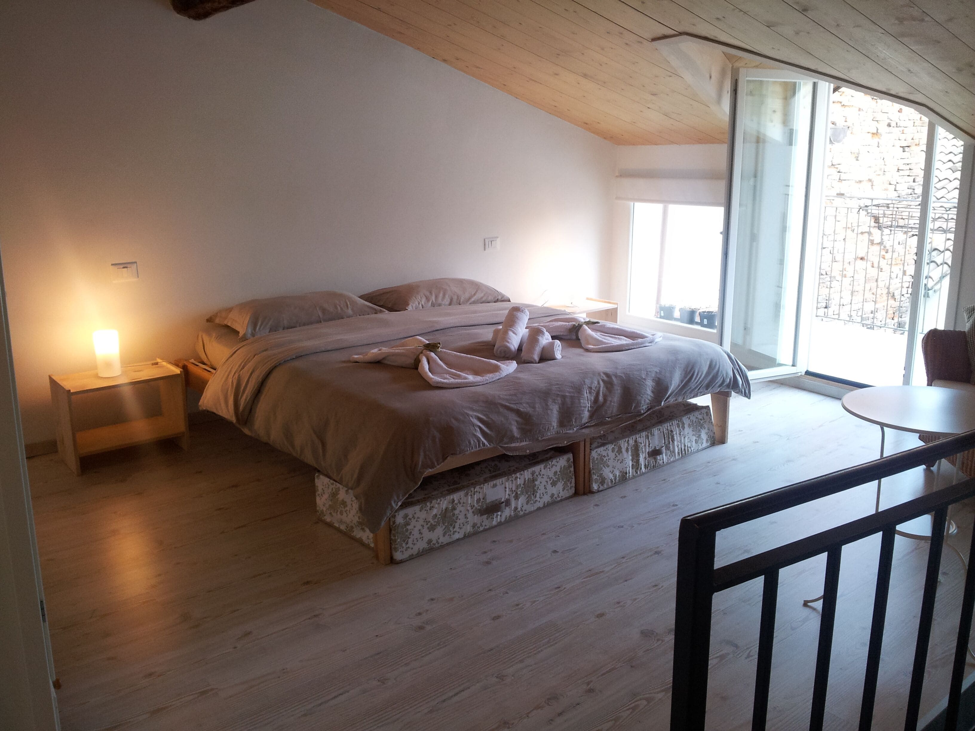 A bedroom with a bed, bedside tables, a small table, and a view of the outside.
