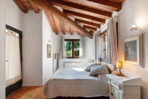 Ein Schlafzimmer mit Holzboden, einem Bett mit floralem Bettzeug, einem Fenster mit Vorhängen und einer Nachttischlampe.