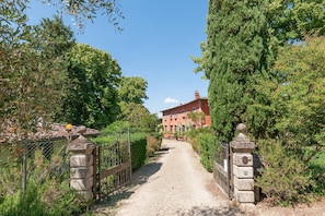 A gravel path leading to a red building with multiple windows, surrounded by greenery and stone gates. - Villa Il Rossignolo Apartment – Private Garden, Shared Pool & Terrace, Wi-Fi (Grassina)