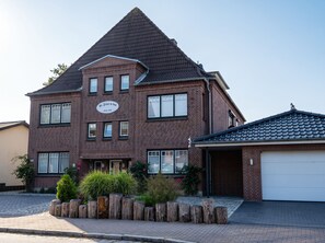A two-story brick house with a gabled roof, a garage, and a small garden area with wooden posts and shrubs. - Gode Stuv: Cozy vacation rental with a balcony (Büsum)