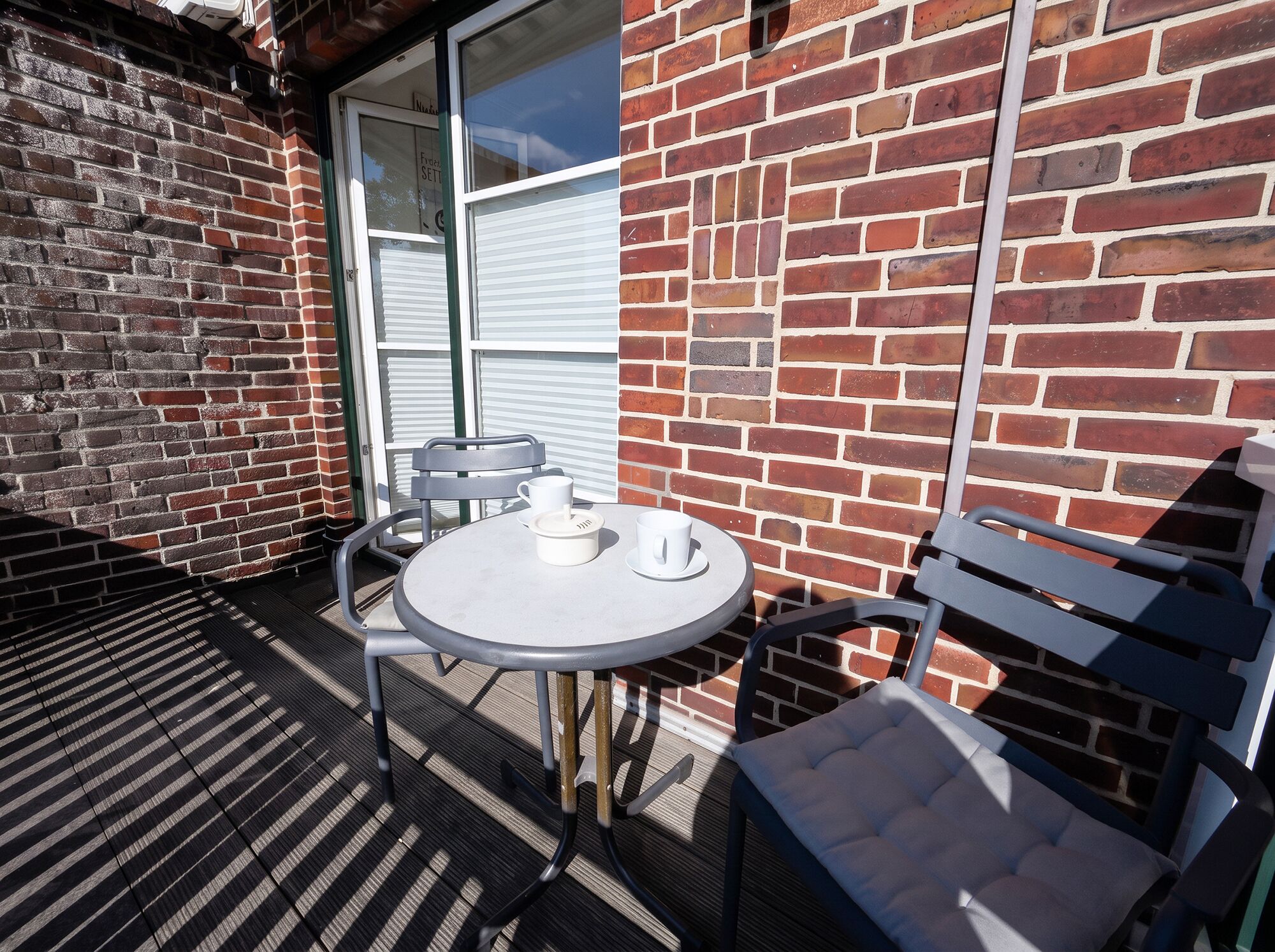 A patio with a round table and two chairs, a brick wall, and a window with blinds.
