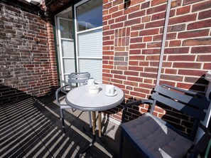 Eine Terrasse mit rundem Tisch und zwei Stühlen, eine Ziegelmauer und ein Fenster mit Rollläden.