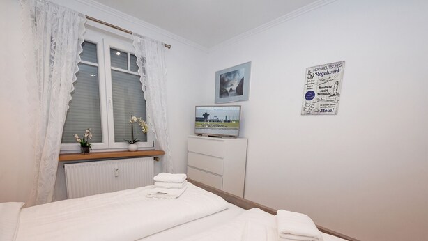 A hotel room with two beds, a window with lace curtains, a radiator, a small table with a plant, a framed picture on the wall, and a sign with text. - Gode Stuv: Cozy vacation rental with a balcony (Büsum)
