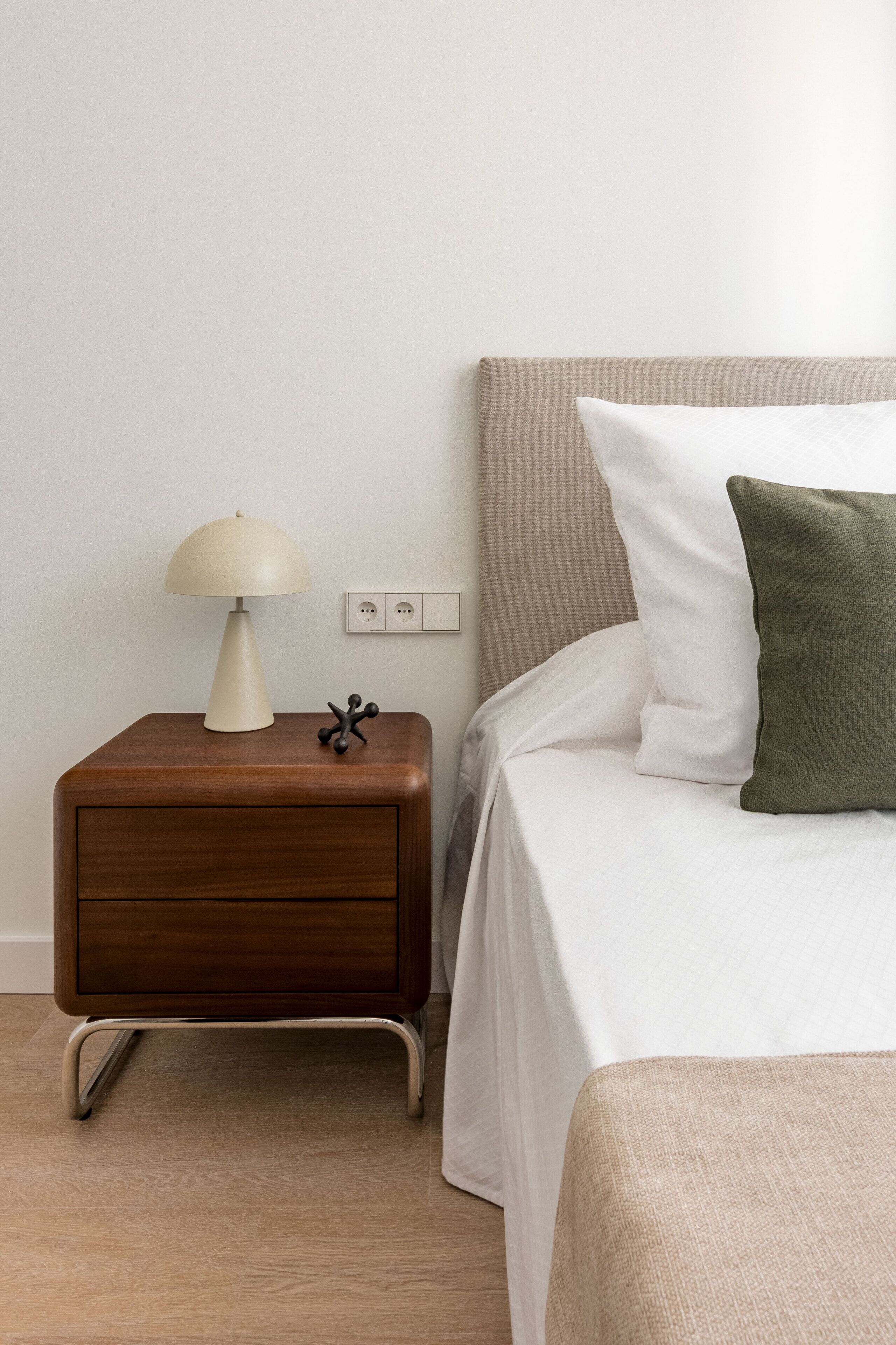 A neatly made bed with white and green pillows, a bedside table with a lamp, and a power outlet on the wall.