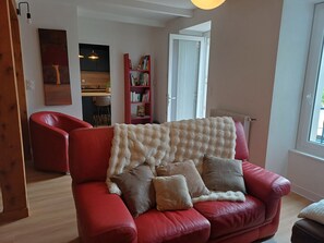 Ein rotes Ledersofa mit flauschiger weißer Decke und Kissen, ein rotes Bücherregal, ein Essbereich aus Holz und eine weiße Tür, die nach draußen führt.