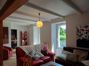 Ein Wohnzimmer mit einem roten Ledersofa, einem Holztisch und einem Bücherregal. Ein Fenster bietet Blick auf Grünfläche.