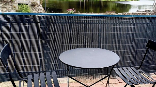 En terrasse med et rundt bord og stole, en stenvæg og udsigt over en sø med træer.