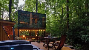 A hot tub, wooden deck, outdoor seating, and a glass enclosure with a forest view. - Mirror house wellness retreat: private sauna, hot tub, spring-fed cold plunge & your own waterfall. Tucked on 35 acres along the French Broad River, minutes from Brevard breweries, Pisgah & DuPont. (Pisgah Forest)