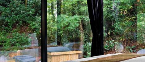 A room with a large window overlooking a forested area, a wooden bench, and a small table.