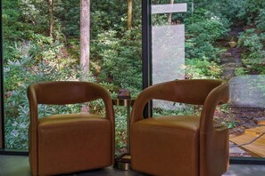 Una habitació amb dues cadires de pell marró, una taula petita rodona i vista a un jardí frondós a través de grans finestres.