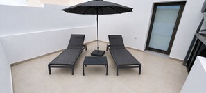 Ein Balkon mit zwei Liegestühlen, einem kleinen Tisch und einem schwarzen Sonnenschirm.