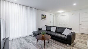 Ett modernt vardagsrum med en svart lädersoffa, ett runt soffbord i trä och en vit gardin.