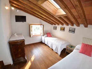 Um quarto com teto inclinado, vigas de madeira, uma cama, uma mesinha e uma janela.