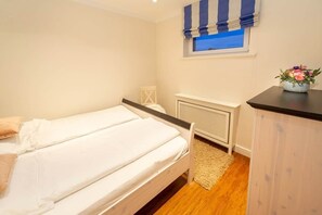 A bedroom with two single beds, a radiator, a window with blue curtains, a small table with a vase of flowers, and a wooden floor. - Feuerstein Family Home, Apartment 4 (Wangerooge)