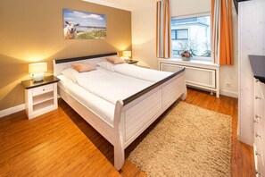 Una cama doble con cabecera, dos mesitas de noche con lámparas, una ventana con cortinas y vista a un edificio.