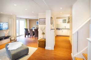 A modern living room with a grey sofa, a dining area with chairs, and a kitchen with white cabinets.