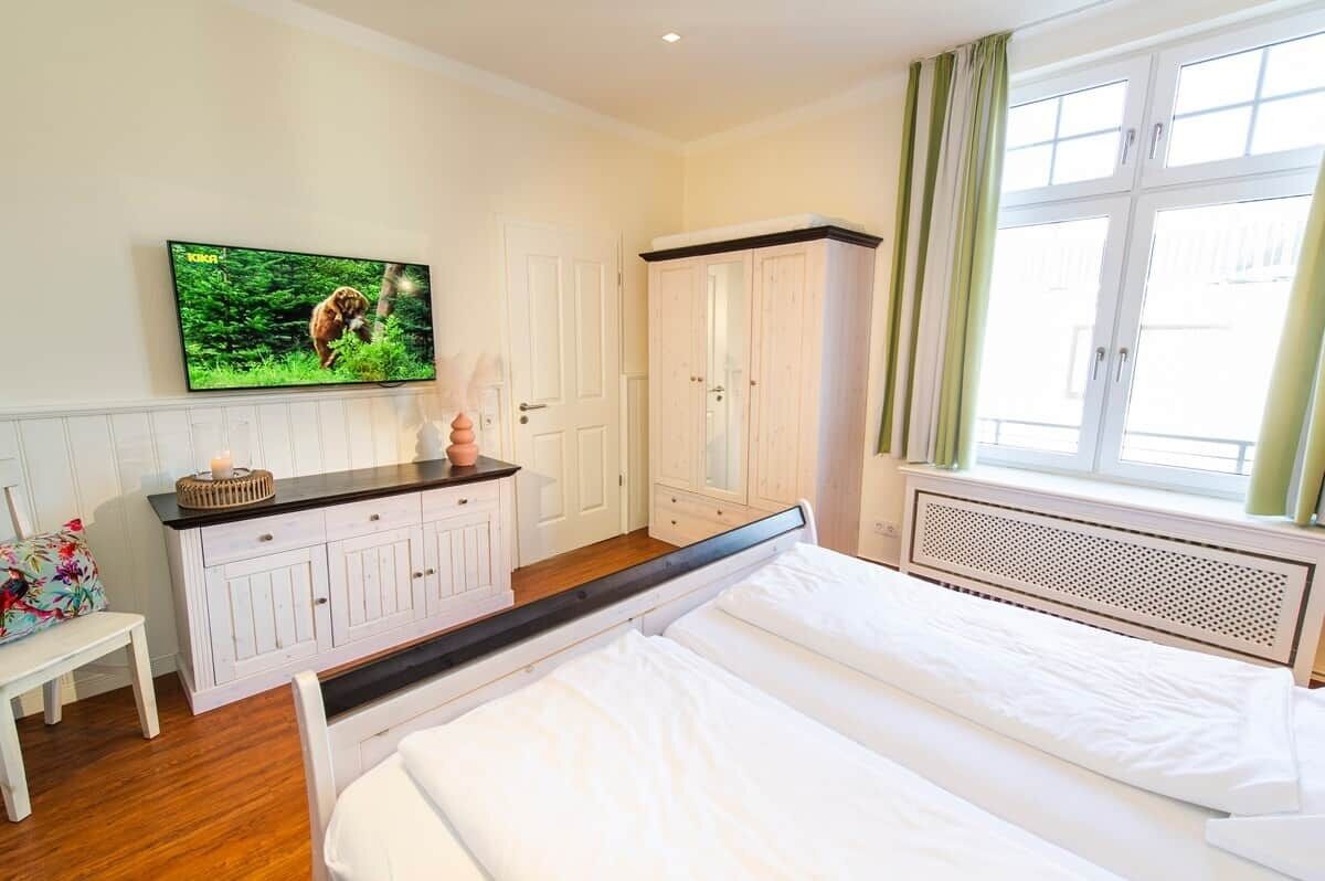Un dormitorio con una cama, un televisor montado en la pared, un armario y una ventana con cortinas.