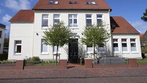 Un bâtiment à deux étages, avec un toit recouvert de tuiles rouges, des murs blancs et une porte d’entrée noire. Deux arbres se trouvent devant le bâtiment, et un vélo est garé sur le côté.