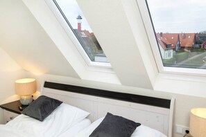 A bedroom with a skylight, a bed with pillows, a bedside table with a lamp, and a view of houses and a lighthouse. - To complete (Wangerooge)