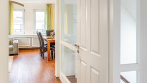 An open door leading to a room with a wooden floor, a dining table with chairs, a sofa, and a radiator. - To complete (Wangerooge)